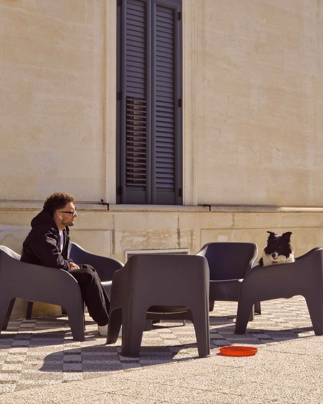 Nicolò Pano seduto al sole su una terrazza, con un cane bianco e nero sulla poltrona di fronte